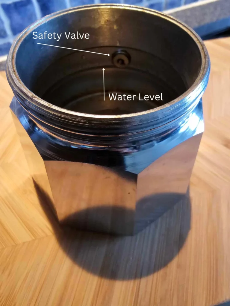 The inside of a moka pot water chamber. White, superimposed, arrows point out the water level and the safety valve.